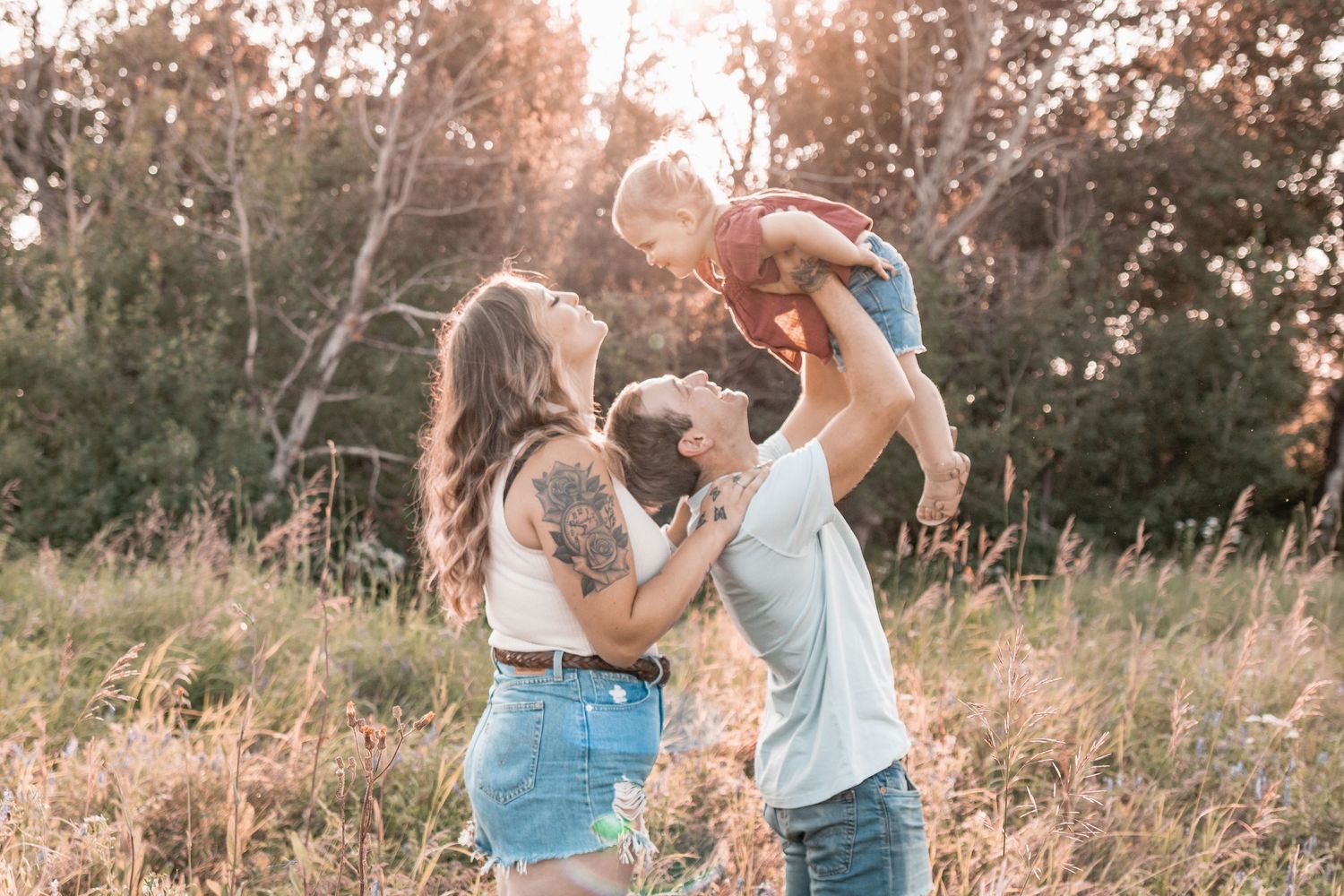 family of three, little girl lifted in air by her dad, leaning back towards mom for a kiss, laughter, outdoor, sunset.