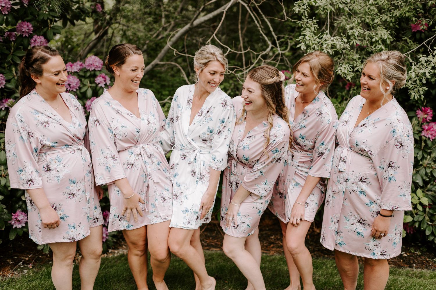 A group of bridesmaids wearing coordinated floral robes laugh together outdoors.