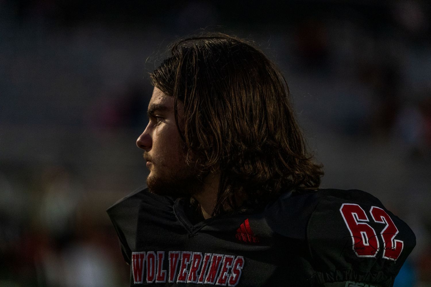 Football player with long hair in black jersey stands on sideline during game.