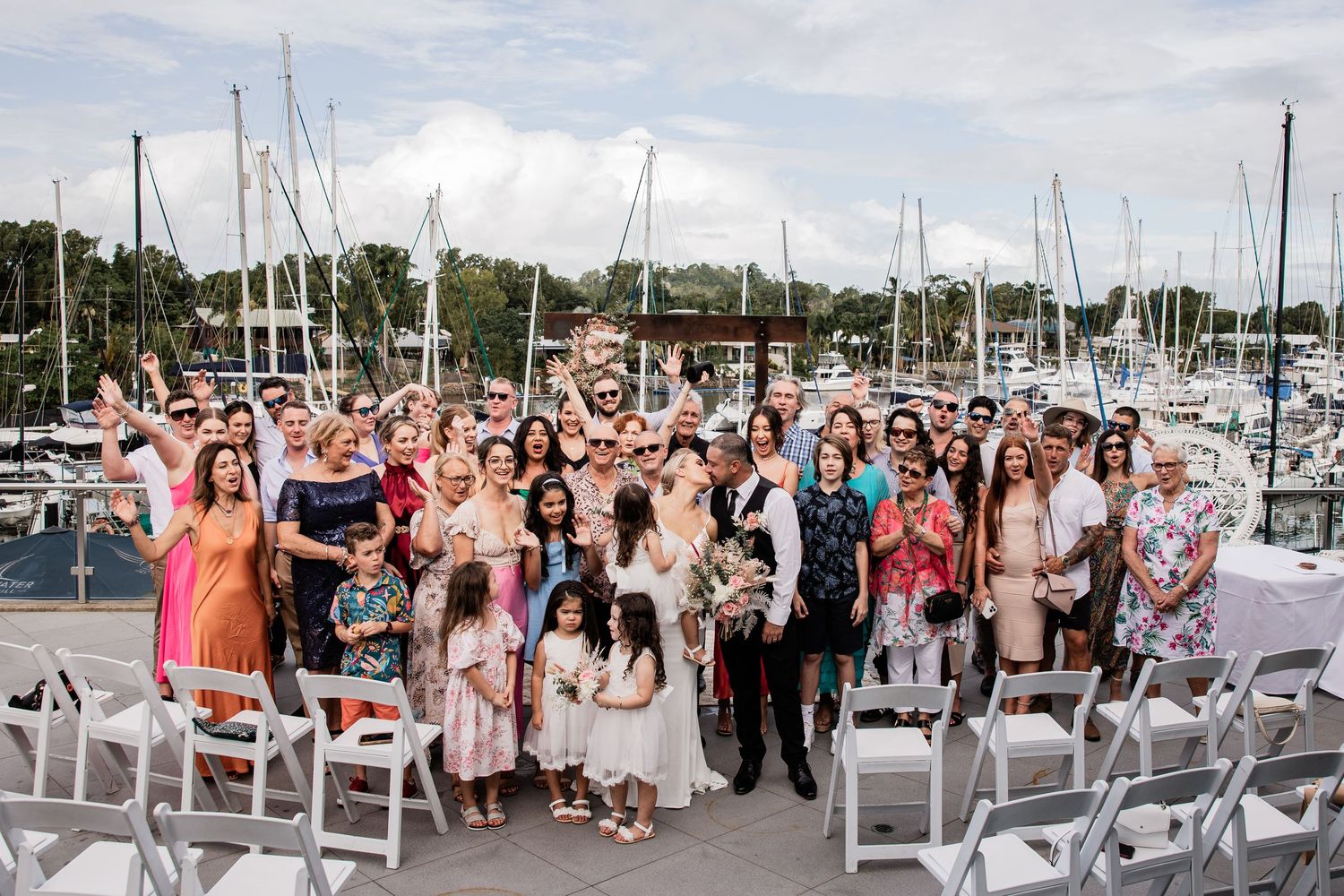 A large wedding group poses together on a marina dock with boats and blue skies in the background.