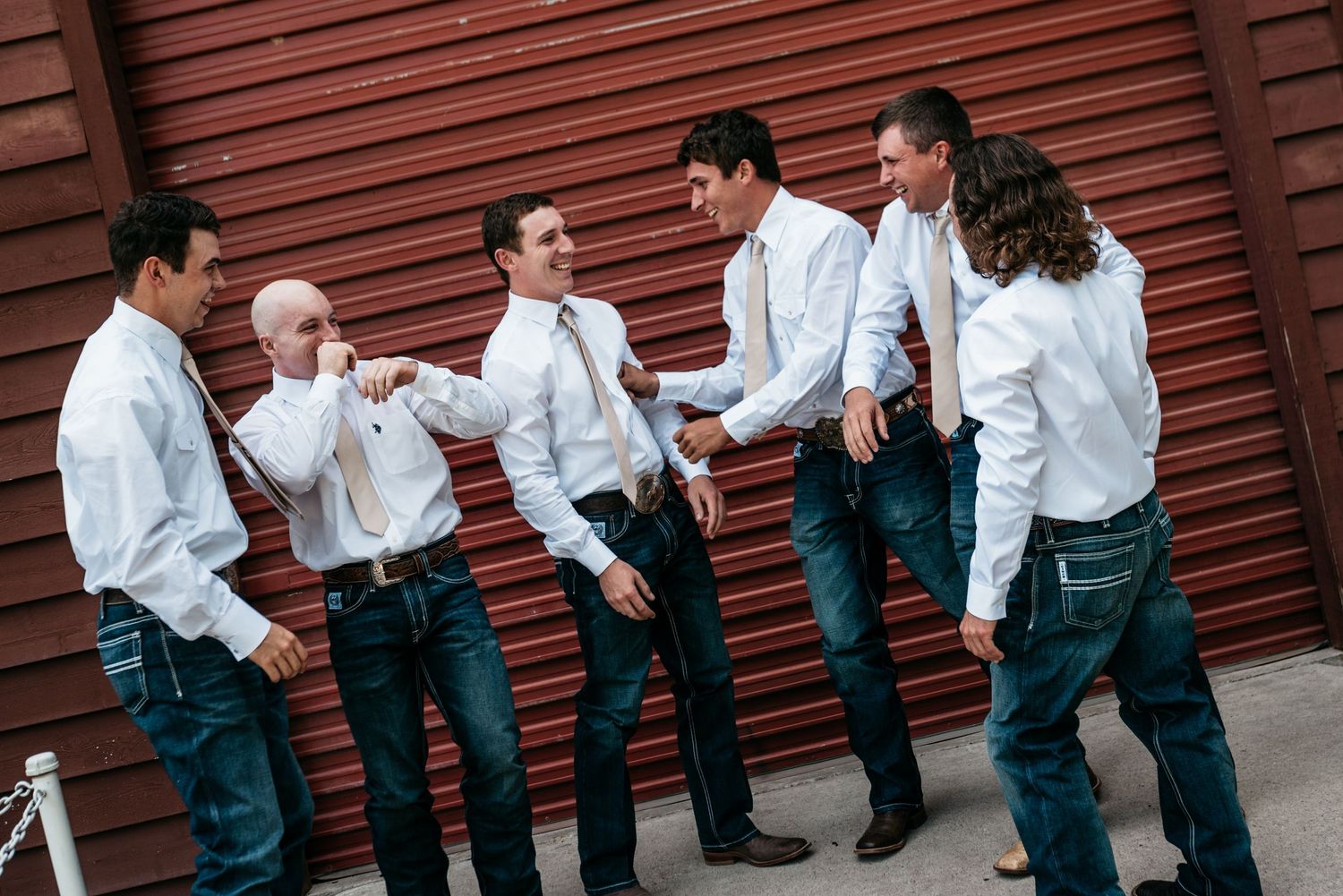 Groomsmen in white shirts and jeans share laughs against rustic red wall.