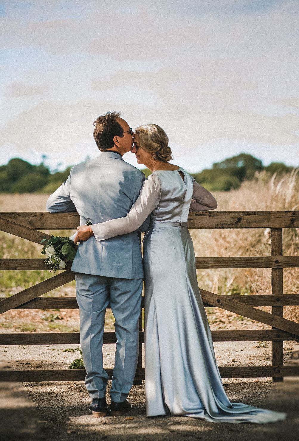 Couple in elegant attire embracing against wooden gate in countryside setting.