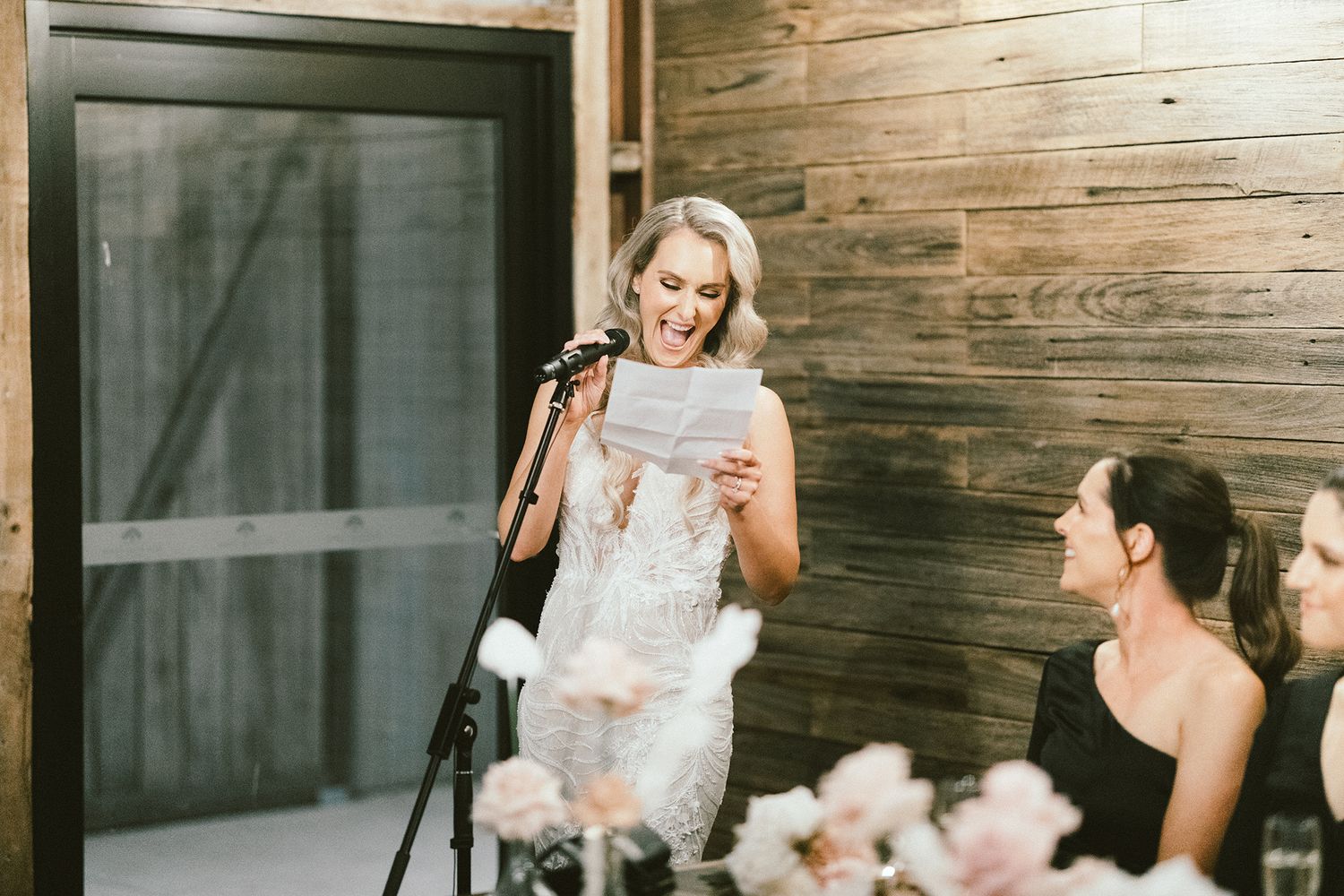 Bridesmaid giving emotional speech at rustic wedding reception with wooden wall backdrop.