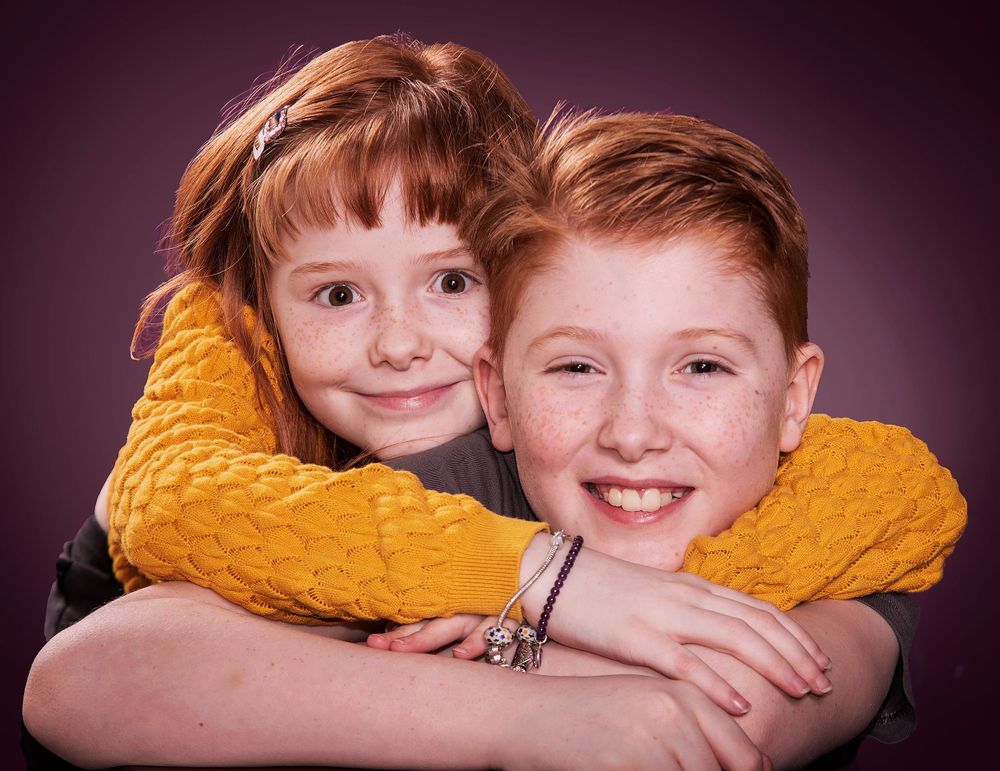 A portrait of two children wearing yellow sweaters against a purple backdrop. for the studio