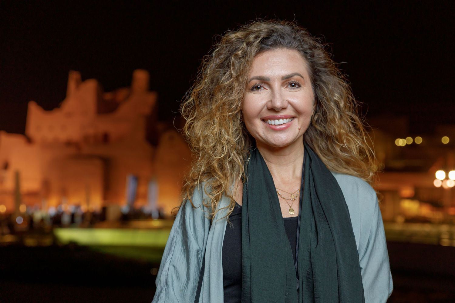 Woman wearing gray jacket and green scarf stands in front of illuminated castle at night.
