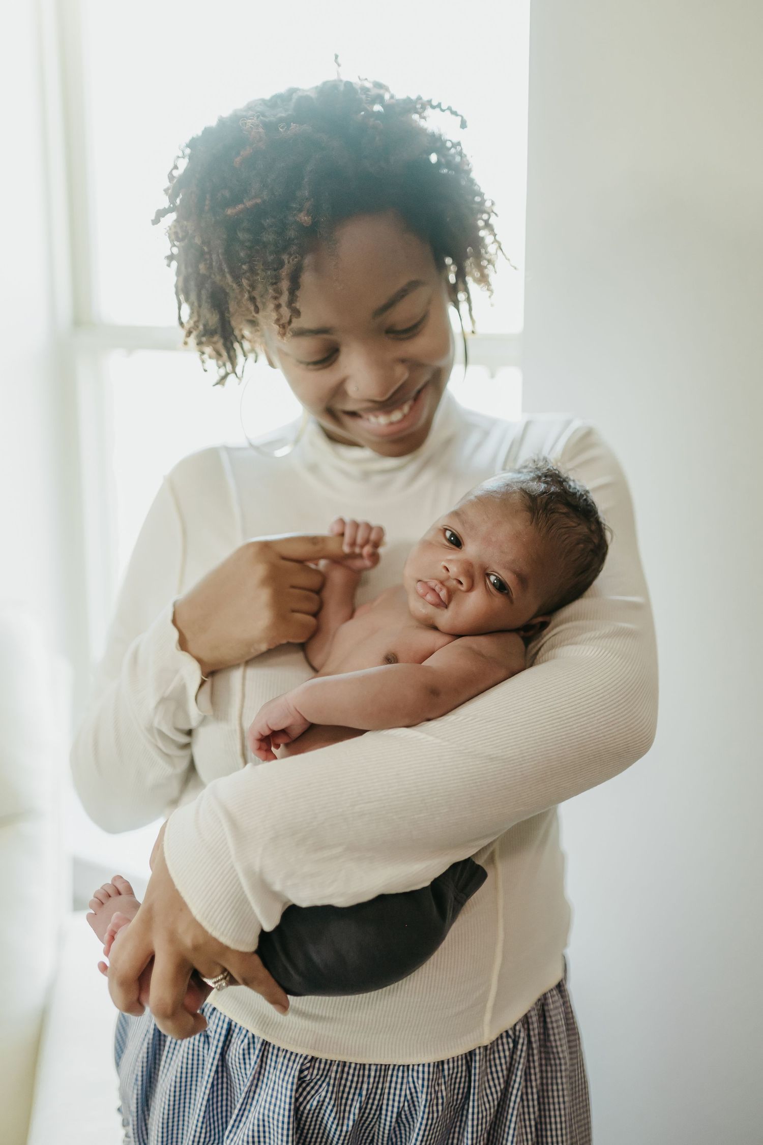 Mother in white sweater holding and smiling at infant against bright window background.