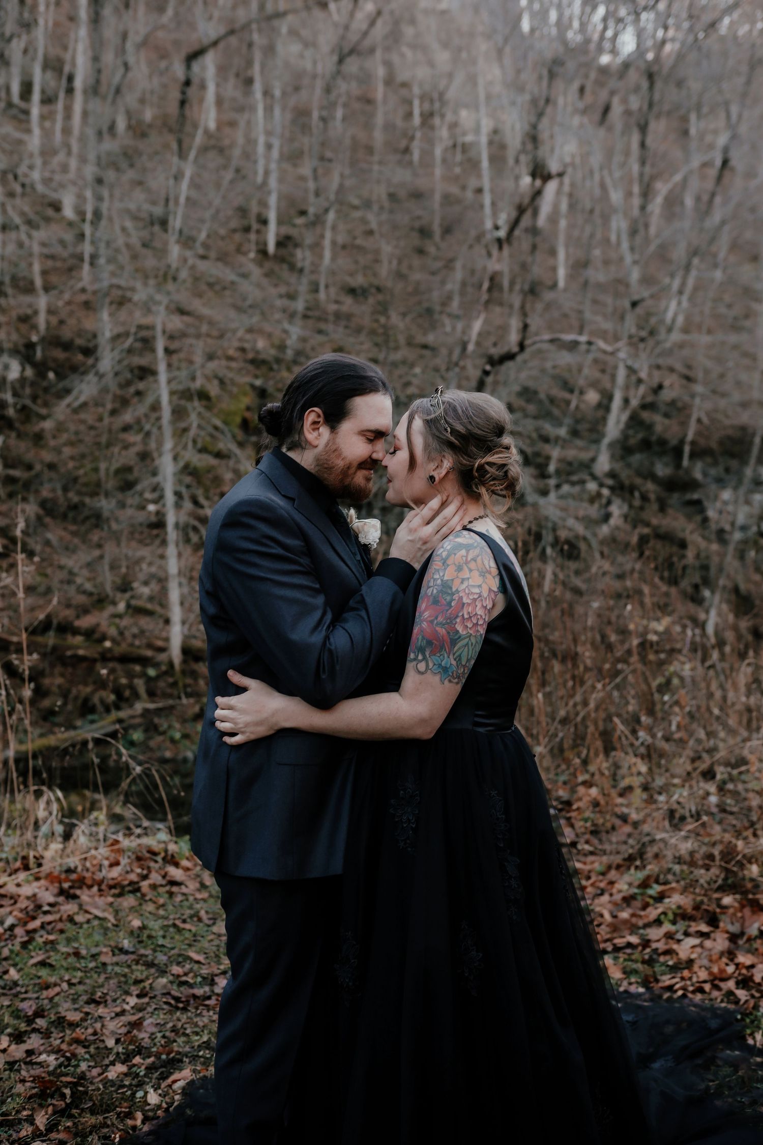 Couple in black wedding attire sharing a romantic kiss in autumn woods.