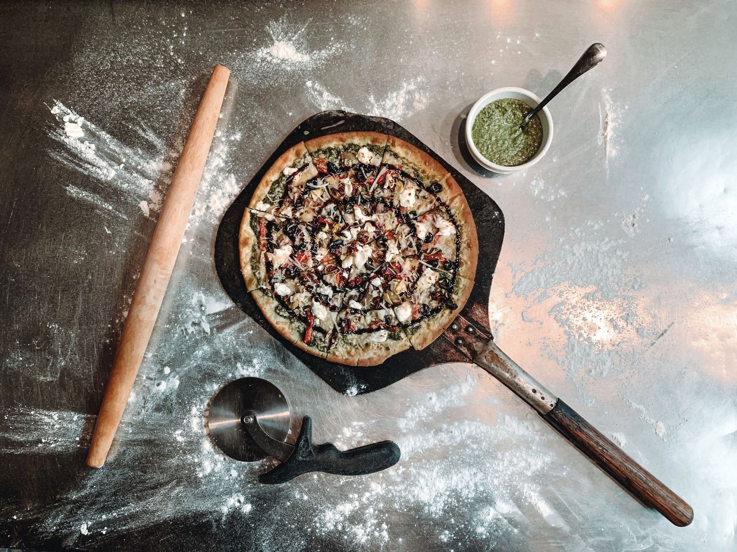 A rustic pizza being prepared in a cast iron skillet with herbs and rolling pin on a floured surface.