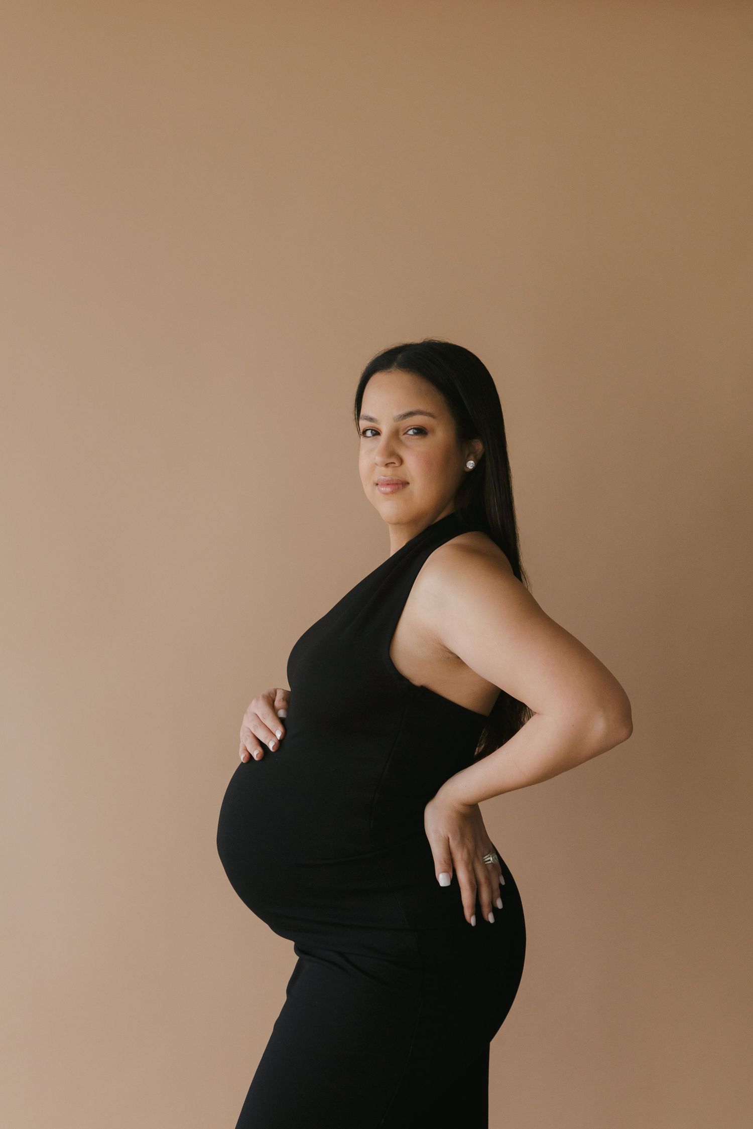 Chic maternity elegance: Paula Ortiz captures a pregnant woman in a sleek black dress against a neutral beige backdrop