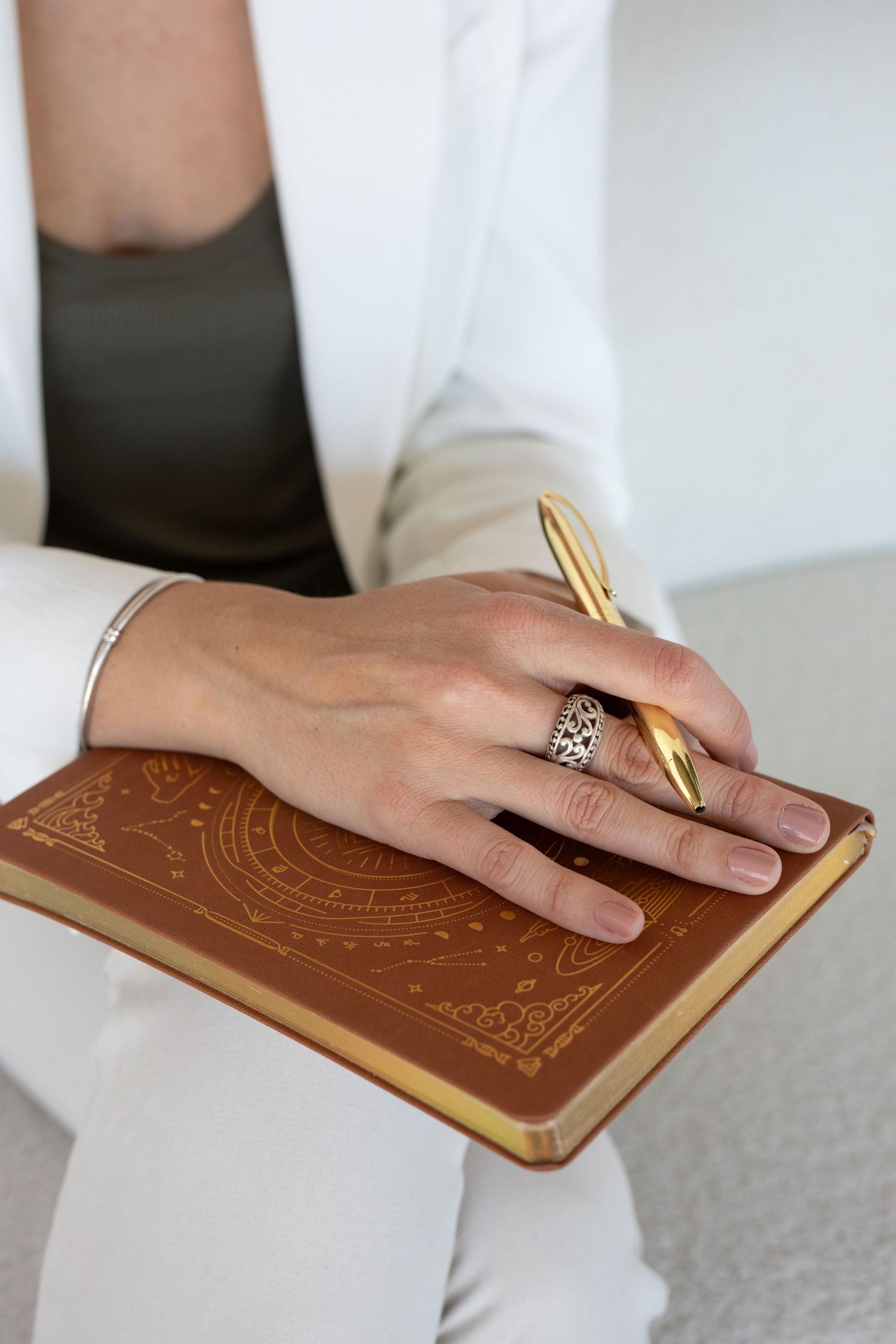 Hand wearing a decorative ring writes in a brown leather journal with a gold pen.