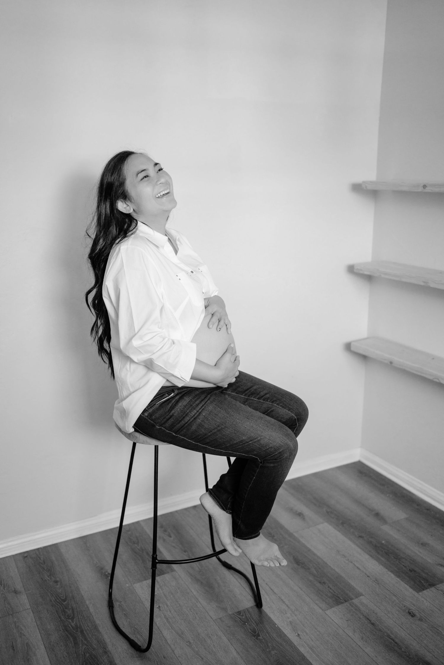 Pregnant woman sitting on a bar stool, head tossed back laughing while cradling her belly. (black and white). Doughman Photography