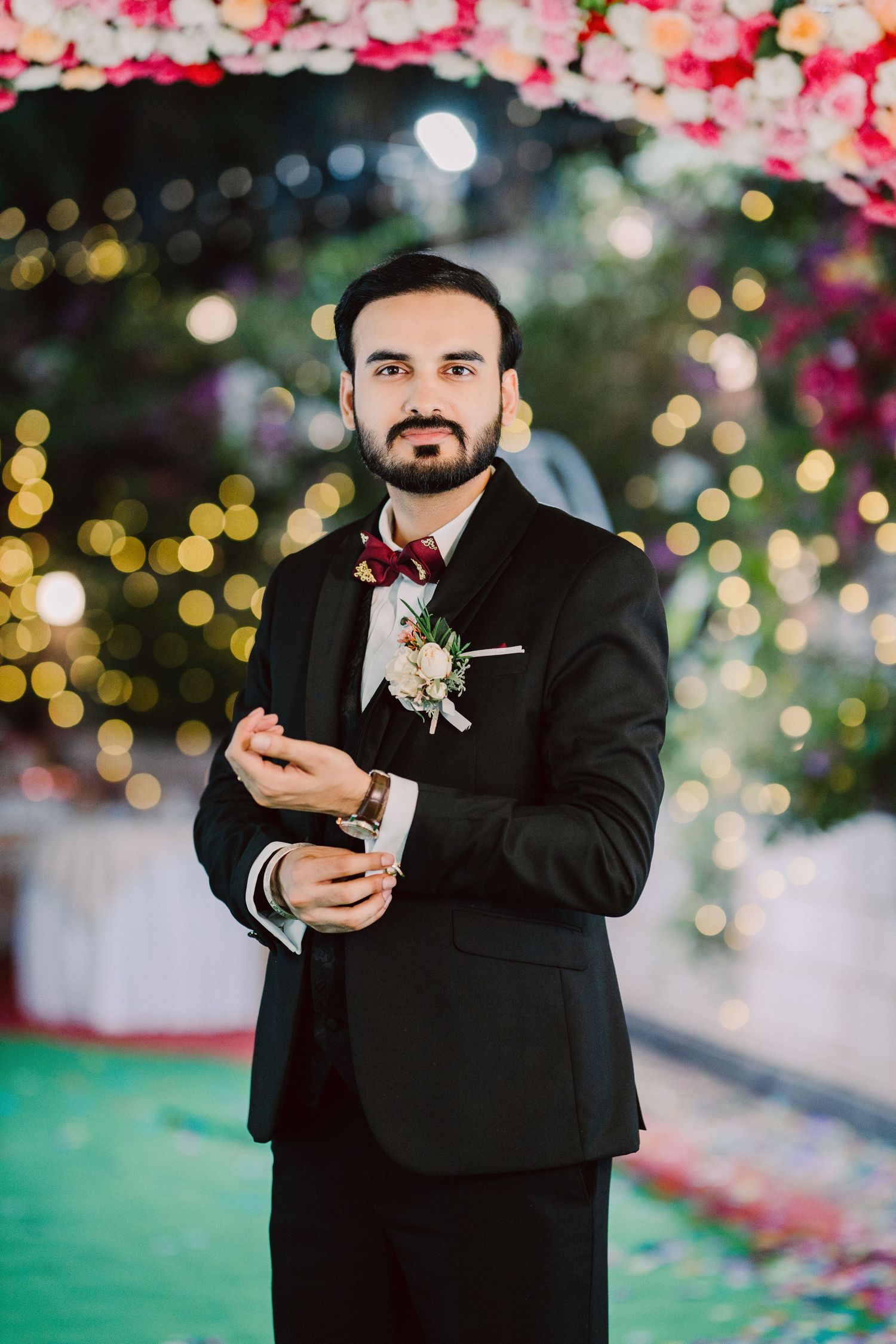 Groom in black tuxedo poses with boutonniere against bokeh background.