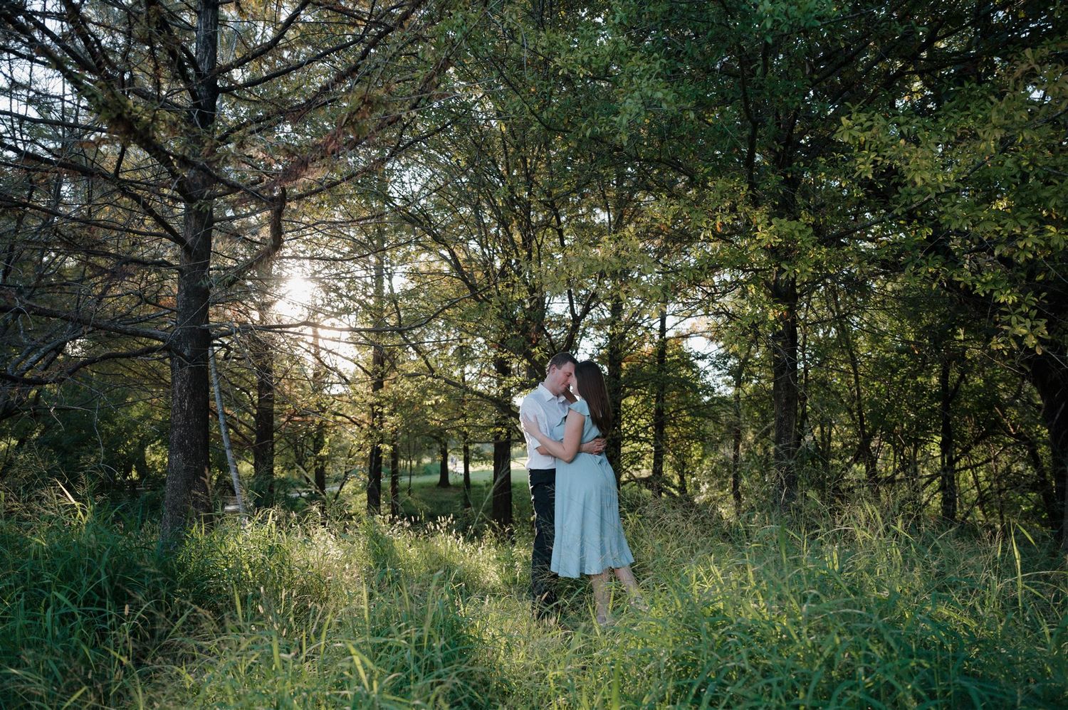 A romantic moment captured in a sunlit forest meadow with tall grass and pine trees at sunset.