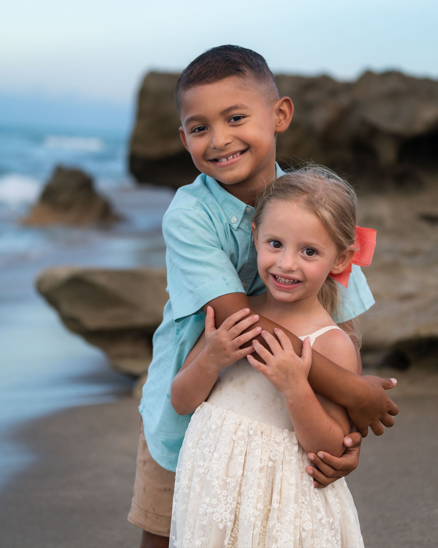 Two children smiling and hugging on a beach with rocky cliffs at sunset.