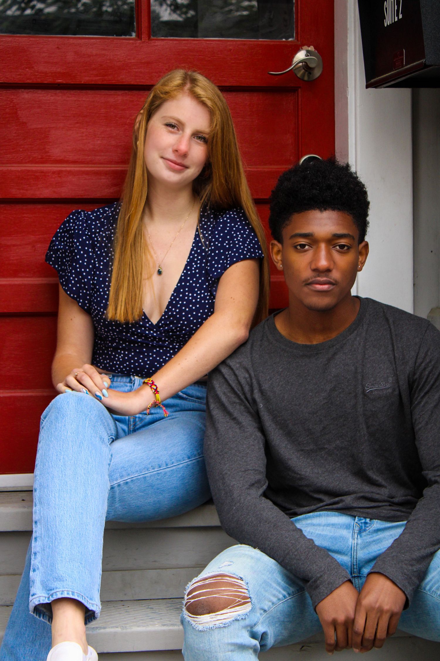 Two young people sit casually on steps in front of a red door wearing jeans and casual tops.