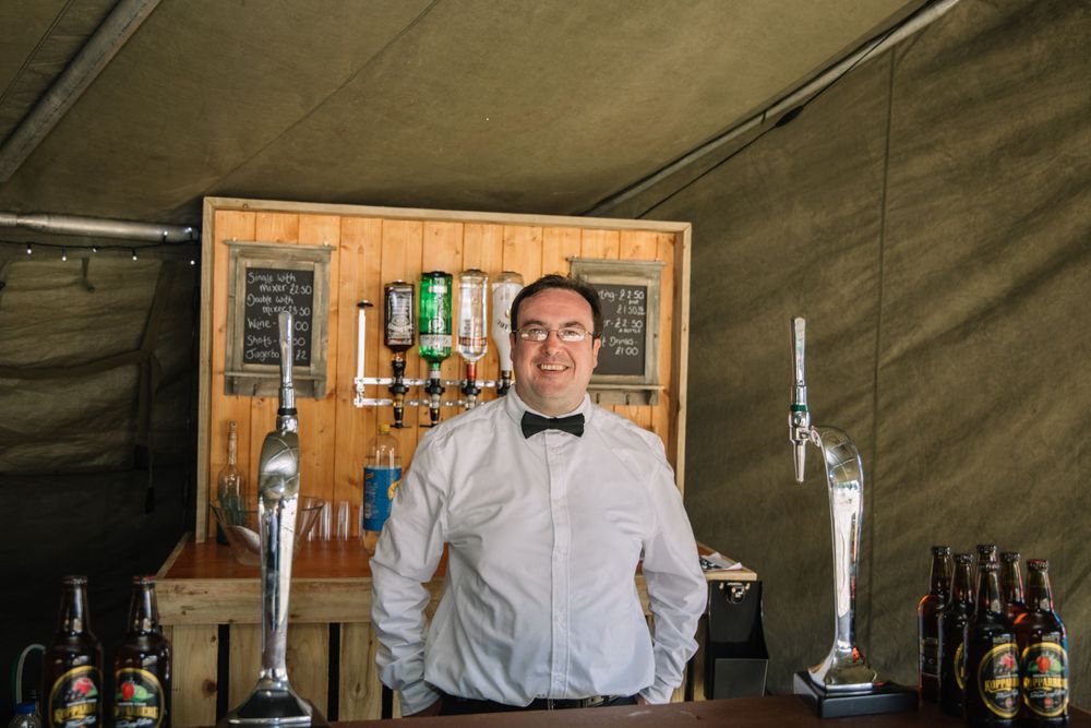 A bartender in bow tie and white shirt stands behind a rustic wooden bar.