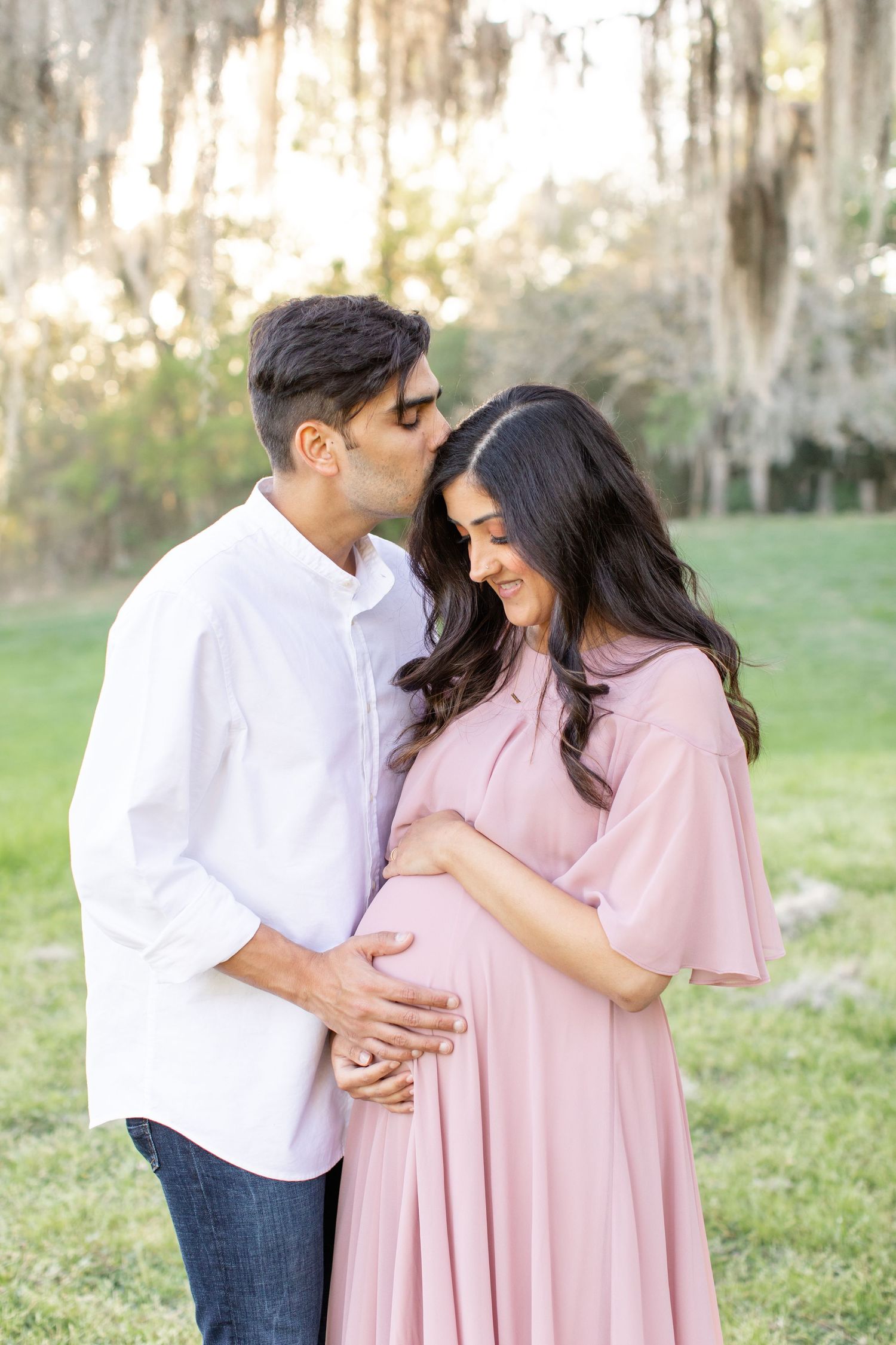 Intimate maternity portrait of couple in coordinated outfits beneath Spanish moss trees.