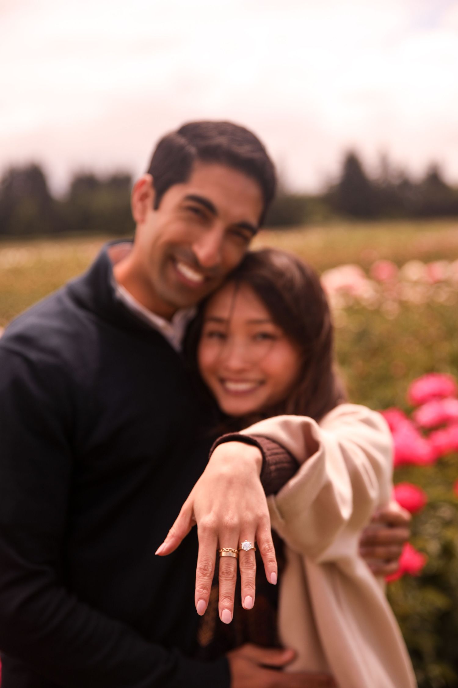 A couple shows off their engagement ring in a field of pink and white flowers.