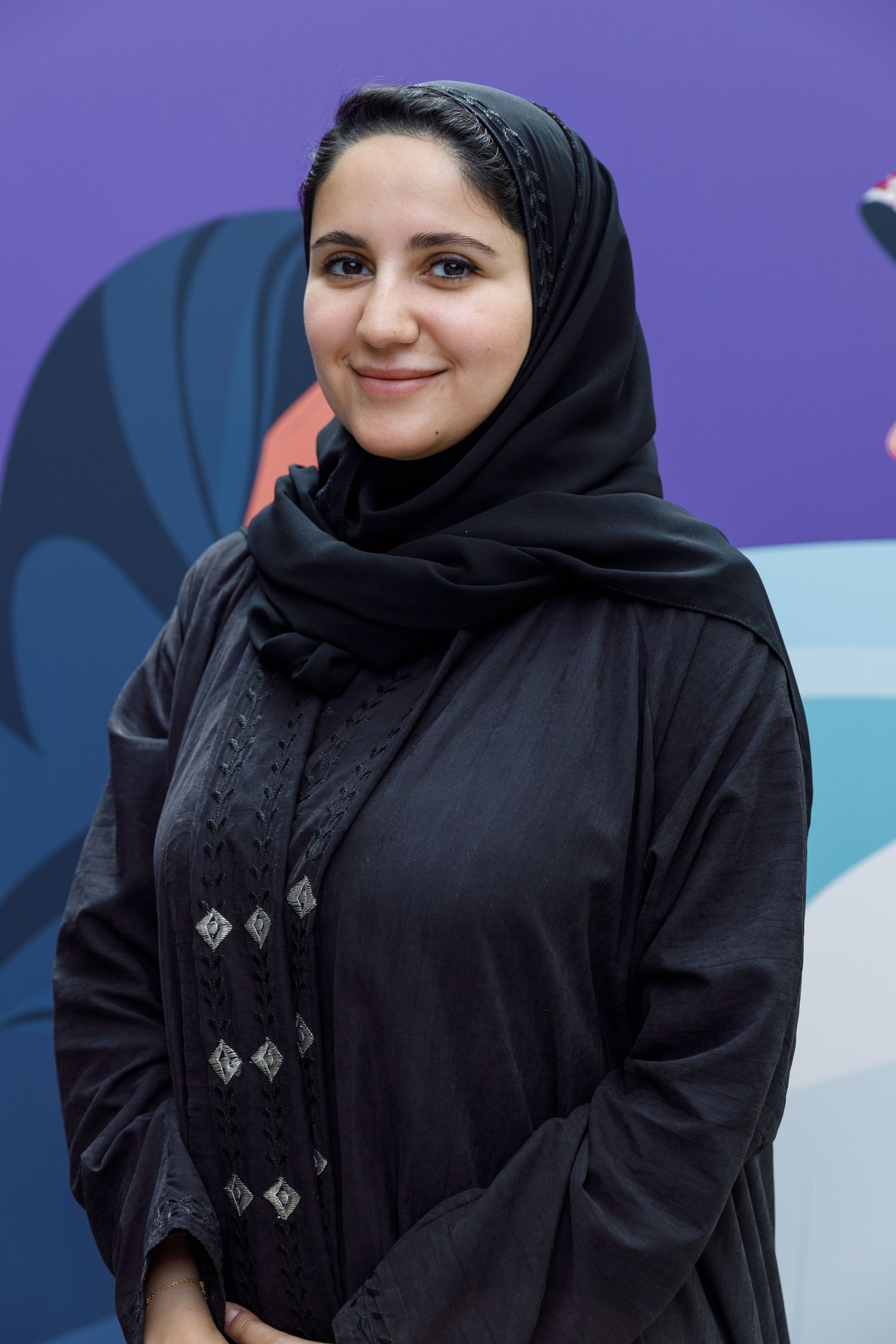 A woman in a black hijab and traditional dress smiling against a colorful background.