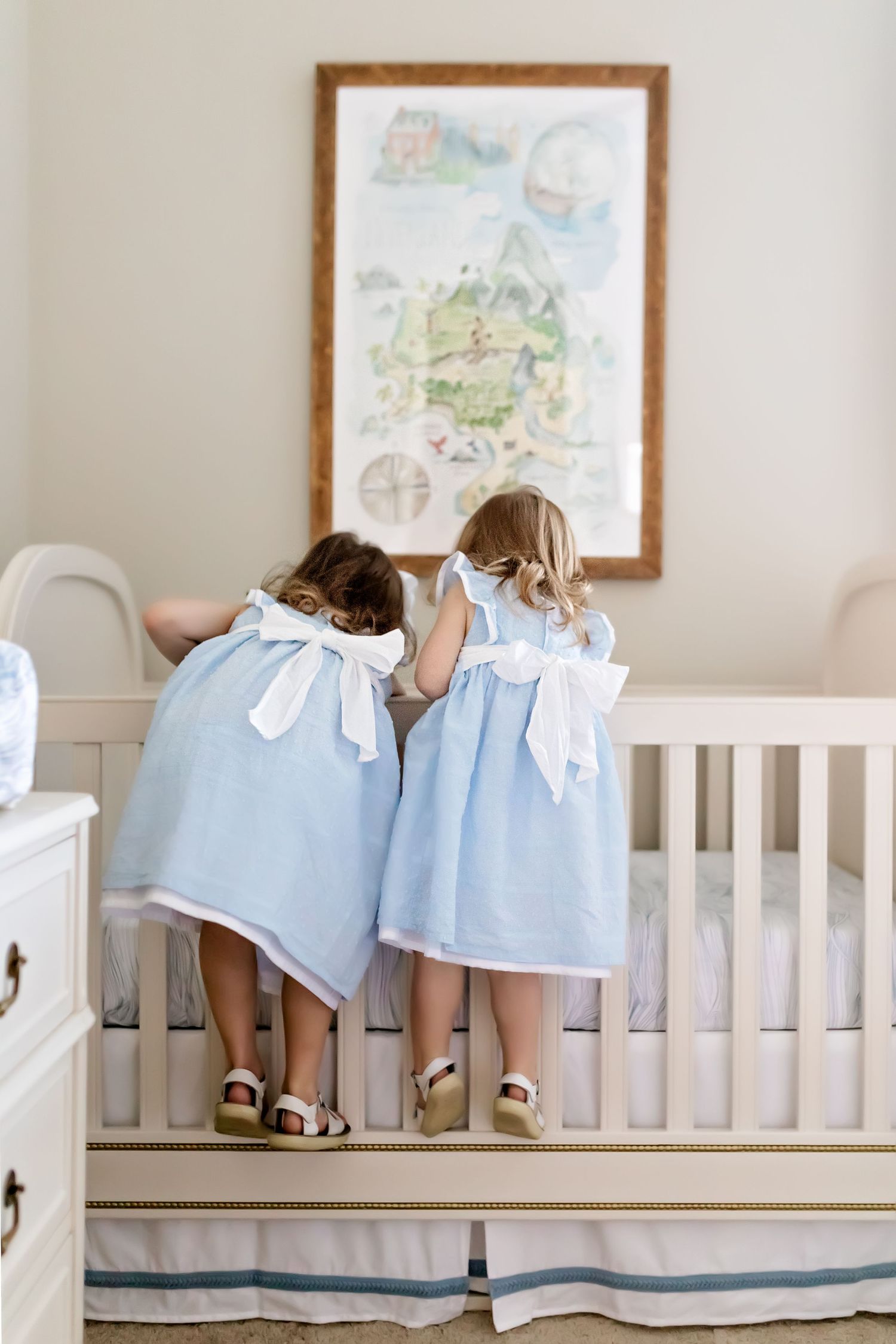 Two young children in matching blue dresses looking at a framed nursery map on the wall.