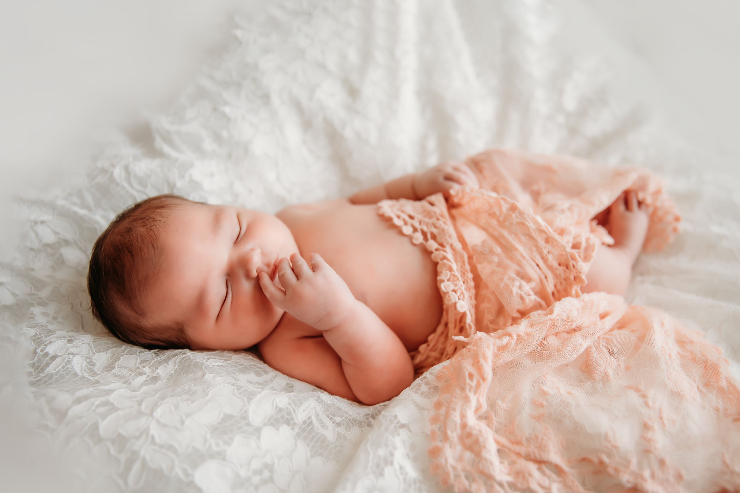 Sleeping newborn wrapped in delicate peach lace on white fur blanket.