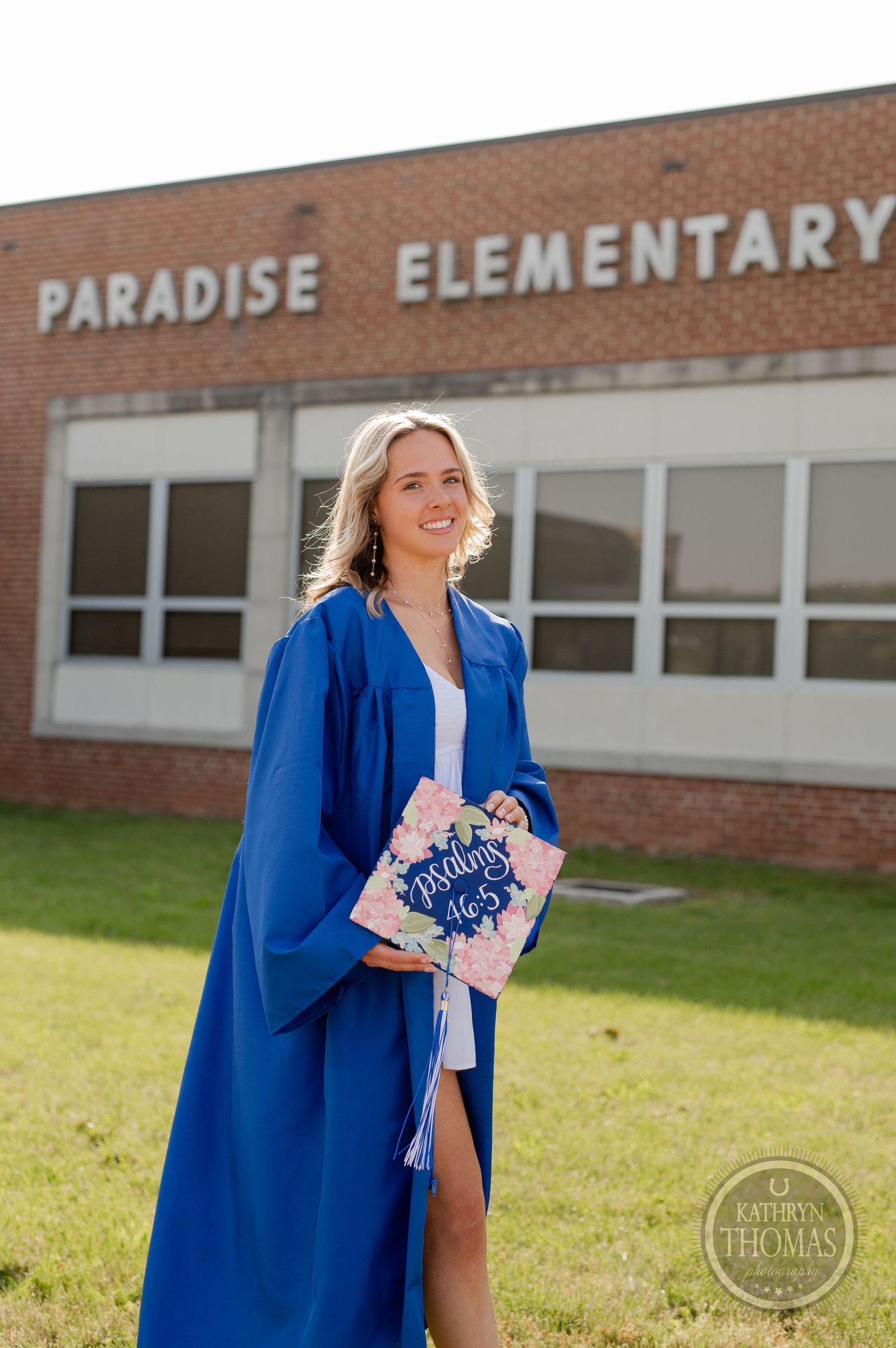 Graduate holds diploma while wearing traditional blue graduation gown at elementary school.