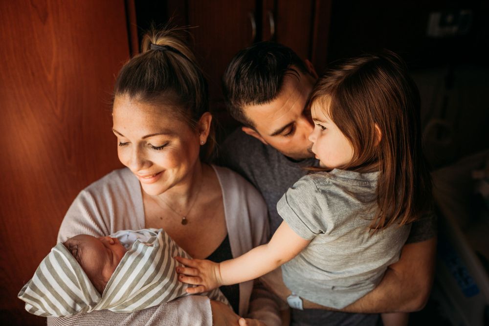 Happy new parents smiling with newborn baby and young daughter by window.