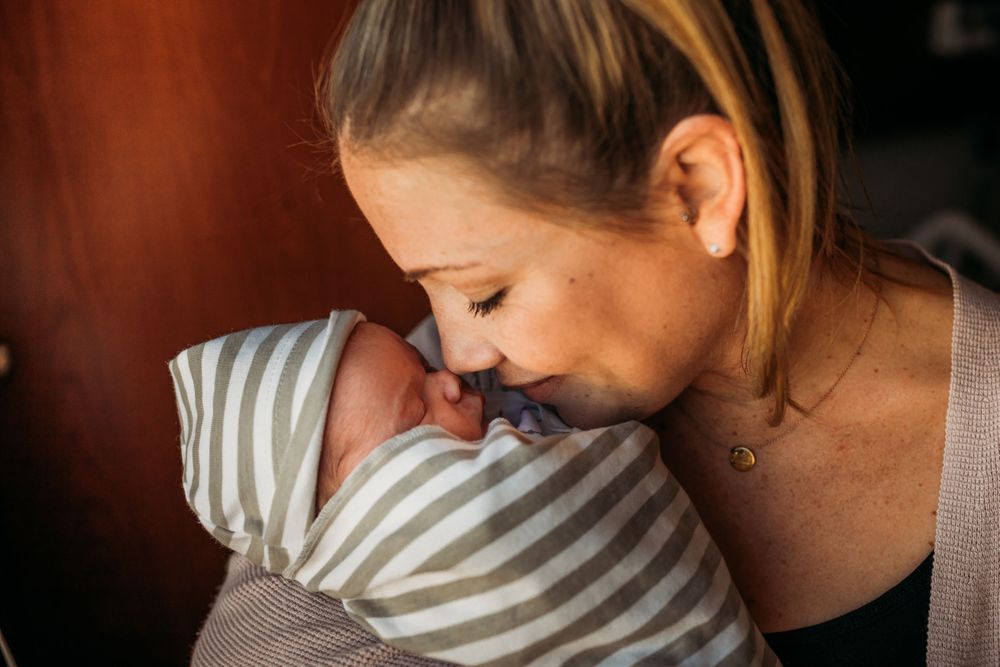 A tender moment as a mother gently kisses her swaddled newborn baby wrapped in a striped blanket.