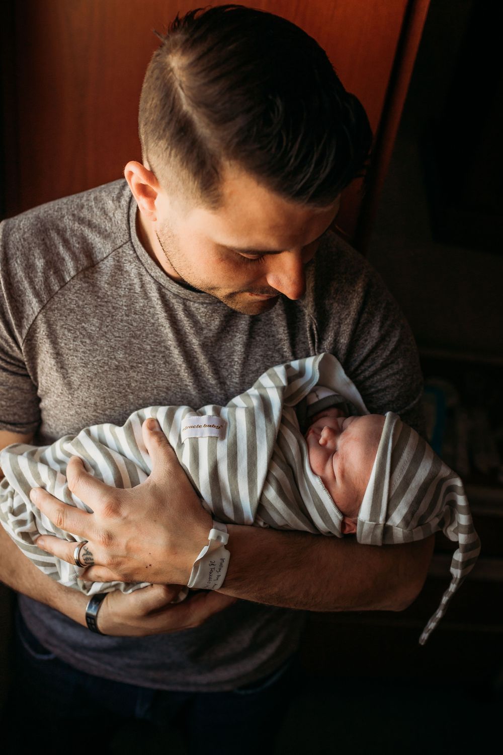 A proud father in a gray shirt carefully cradles his sleeping newborn baby in a striped swaddle.