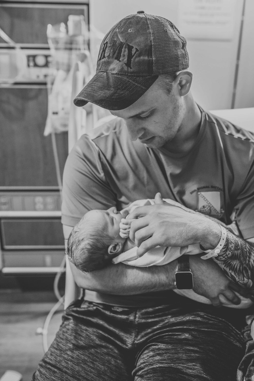 New father in Army cap tenderly holding his newborn baby in hospital room.
