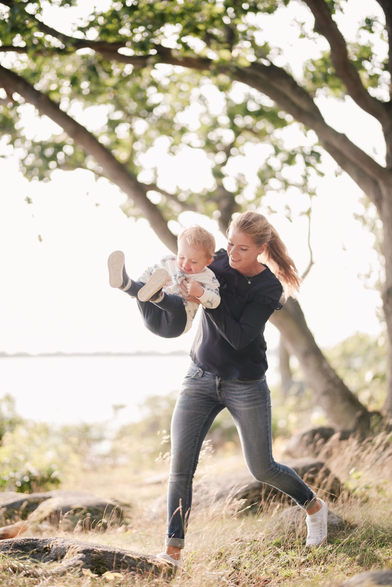 Playful scene by the water with someone swinging another against a backdrop of trees.