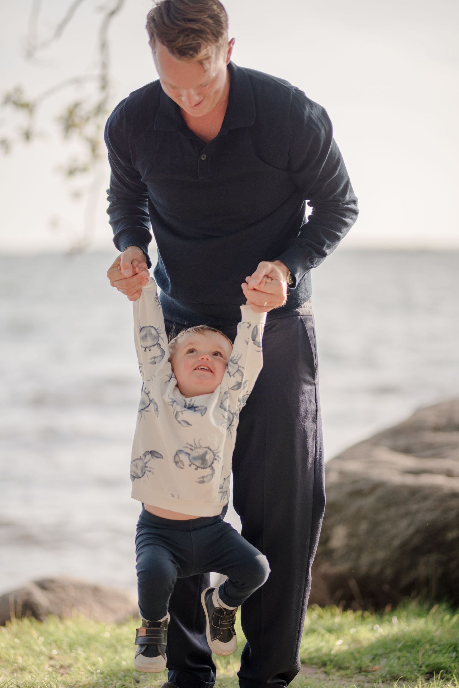 Adult holding young child's hands while playing outdoors near the ocean.