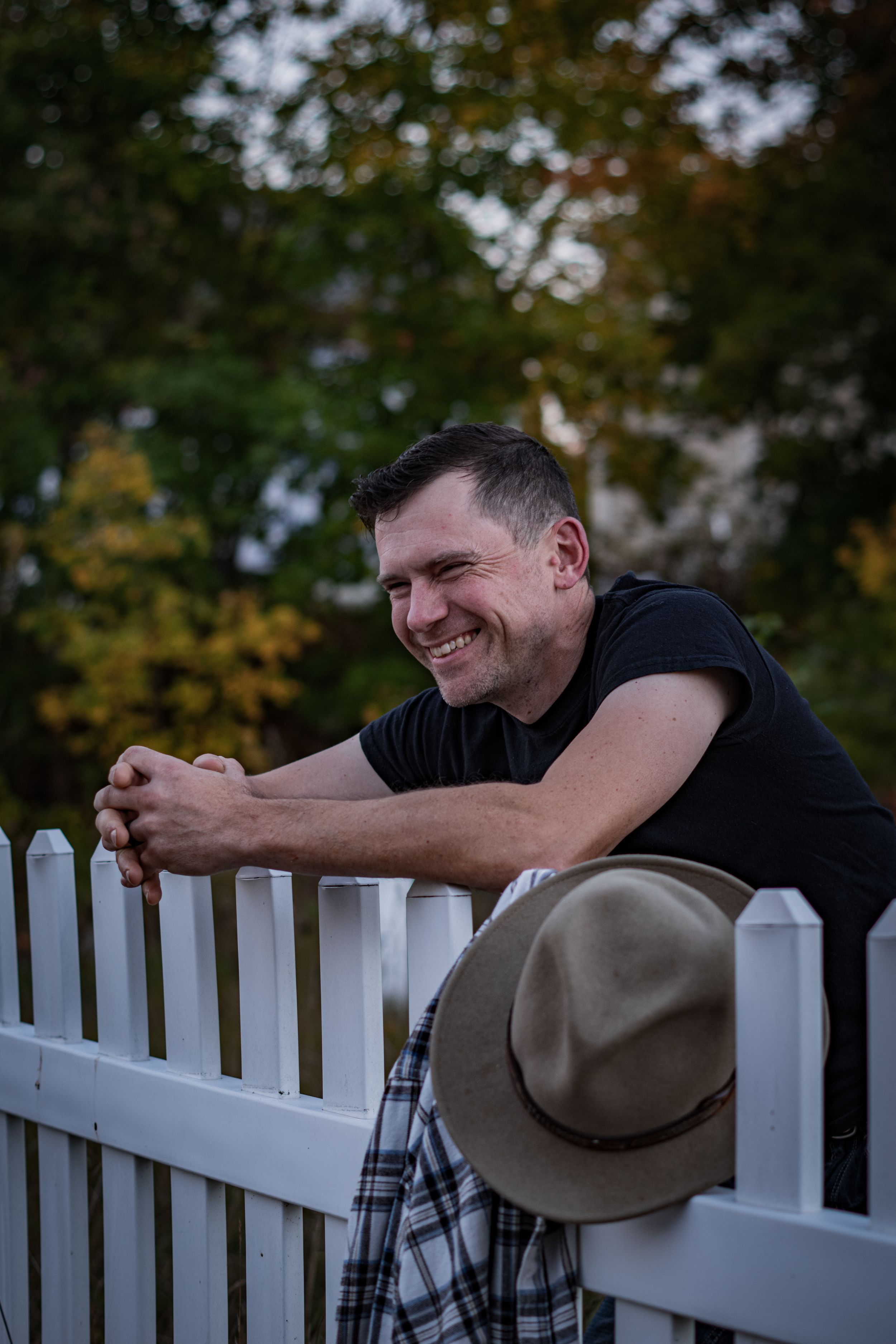 Smiling man leaning on white picket fence with hat hanging nearby, surrounded by autumn trees.