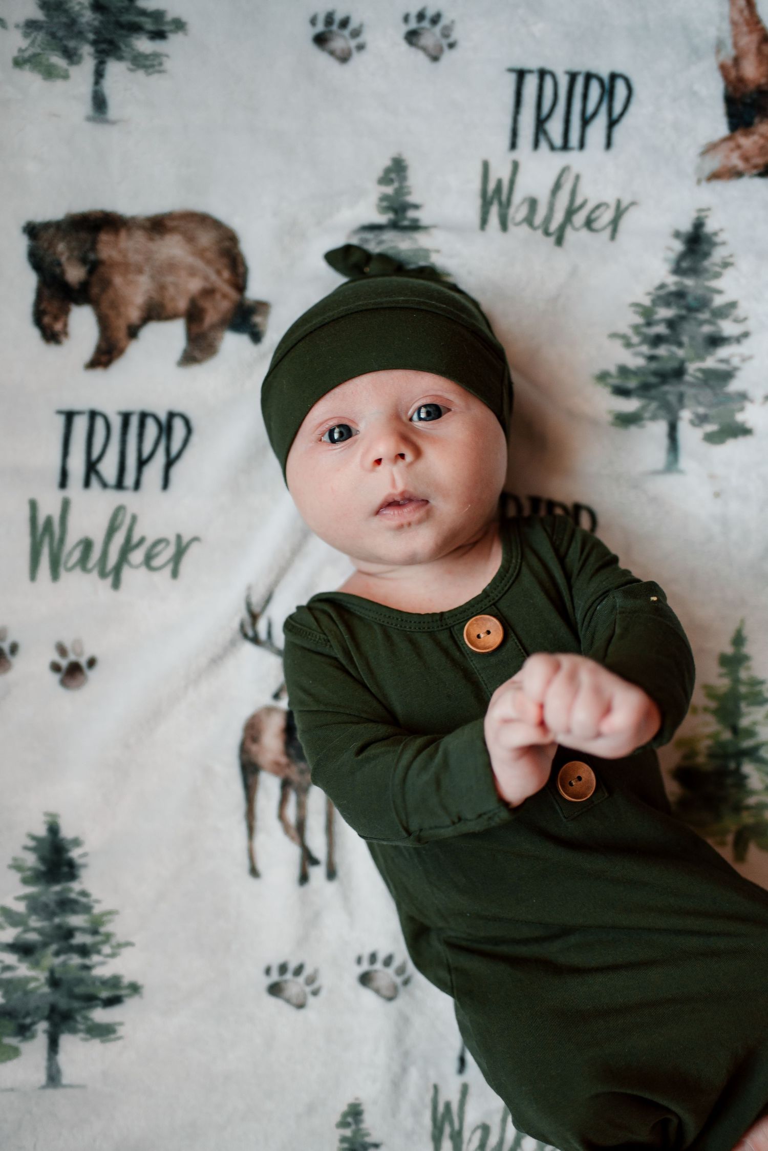 Baby wearing dark green outfit lying on woodland-themed blanket with pine tree and animal prints.