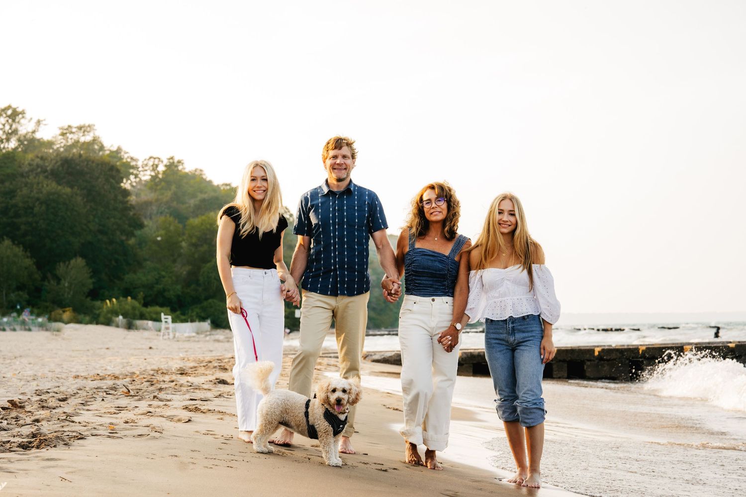 Portrait of family of 4 with dog walking on shore with warm-lit trees in background - shot by Milwaukee family photographer Rachel Dickman.