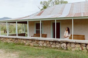 A rustic farmhouse with a stone foundation and metal roof sits against a mountainous backdrop.