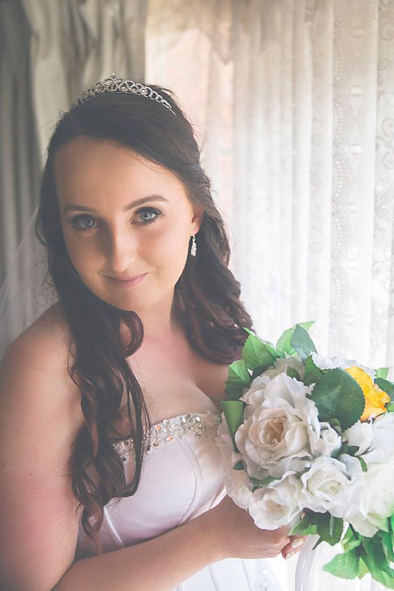 Bride in white dress with tiara poses with white roses against light backdrop.