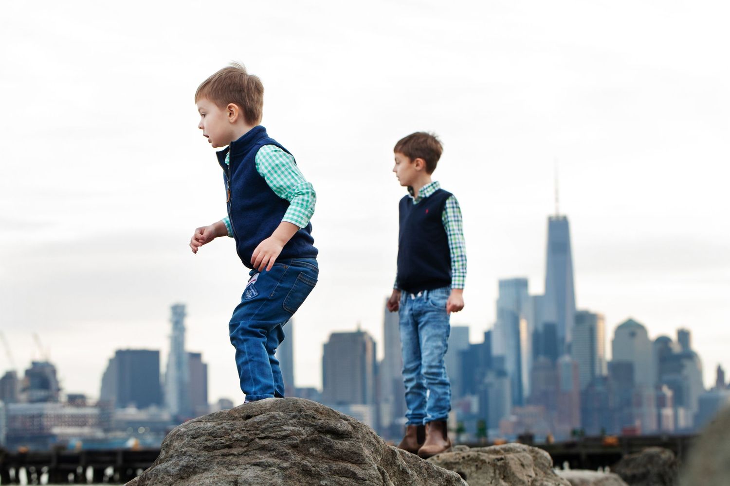 Two children standing on rocks with NYC skyline visible in distance.