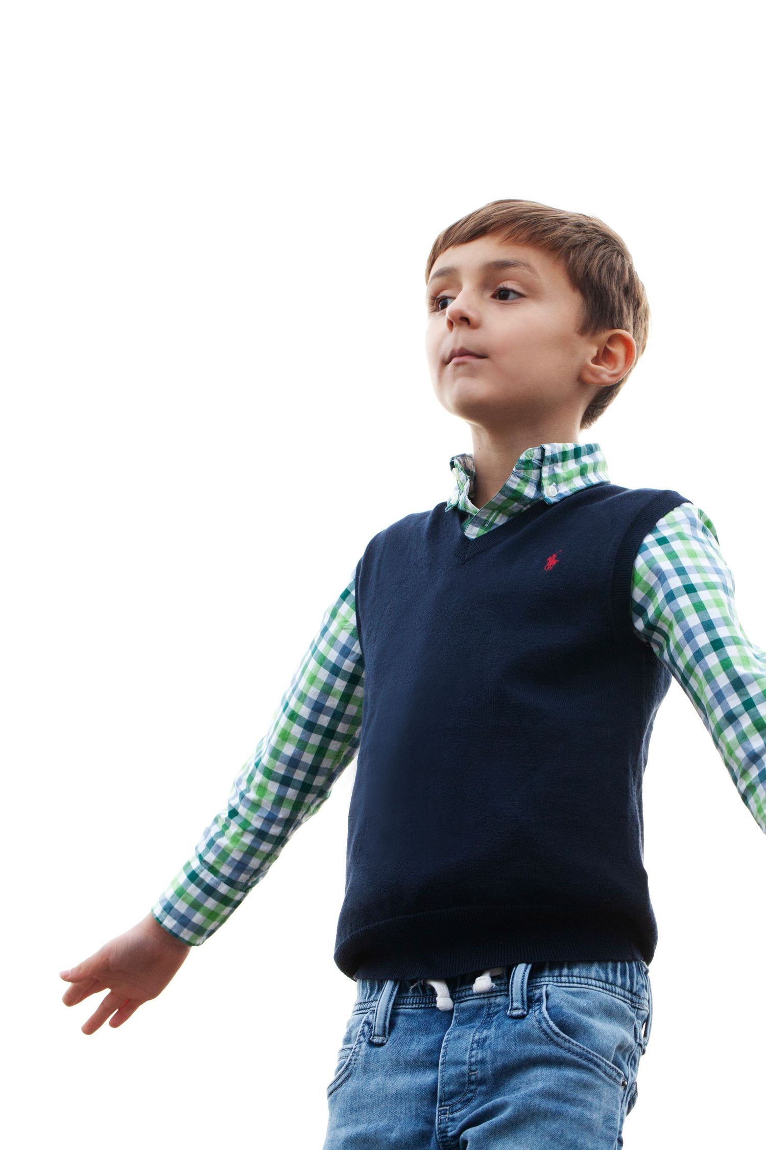 Child in navy vest and gingham shirt looking up against white background.