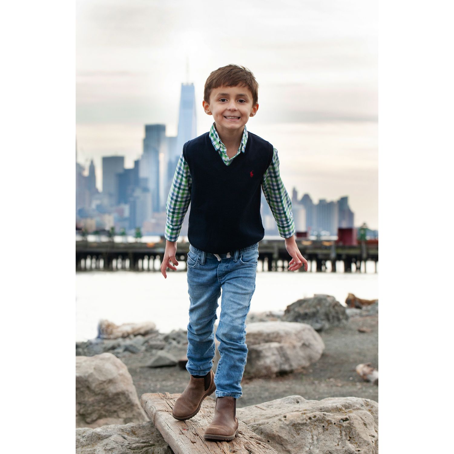 Child in navy vest and gingham shirt stands on driftwood with Manhattan skyline visible.