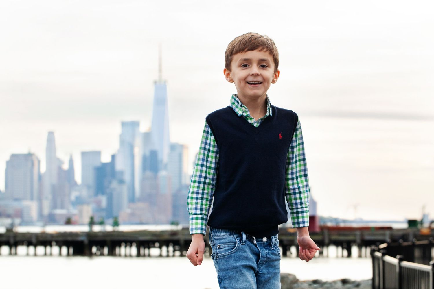 Child in checkered shirt and navy vest smiling with Manhattan skyline in background.