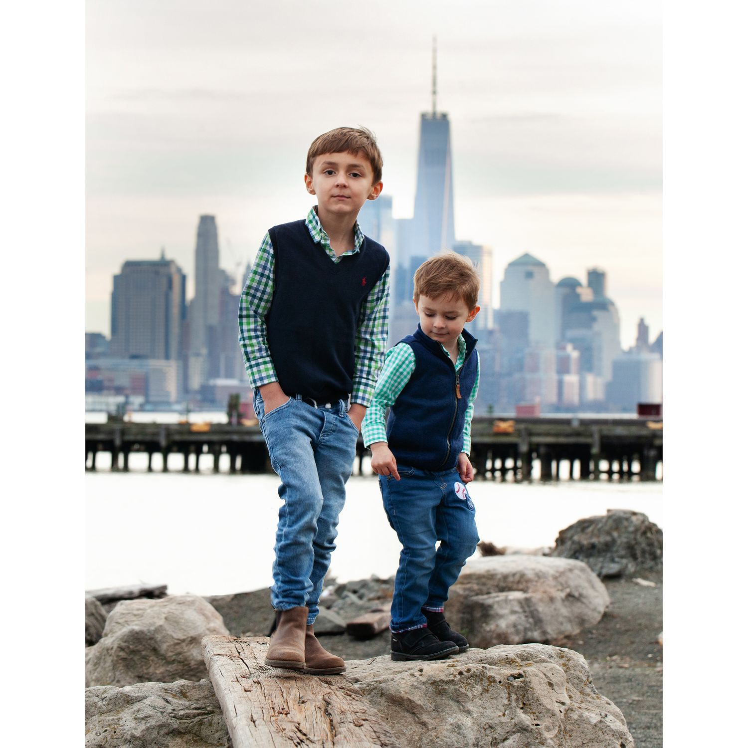 Two children in matching outfits stand on rocks with Manhattan skyline behind them.