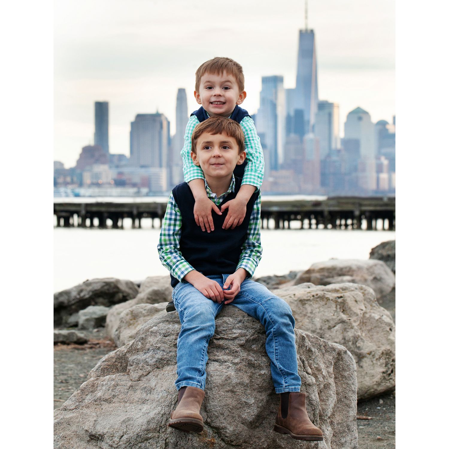 Two kids in coordinating blue vests and gingham shirts pose on rocks with NYC backdrop.