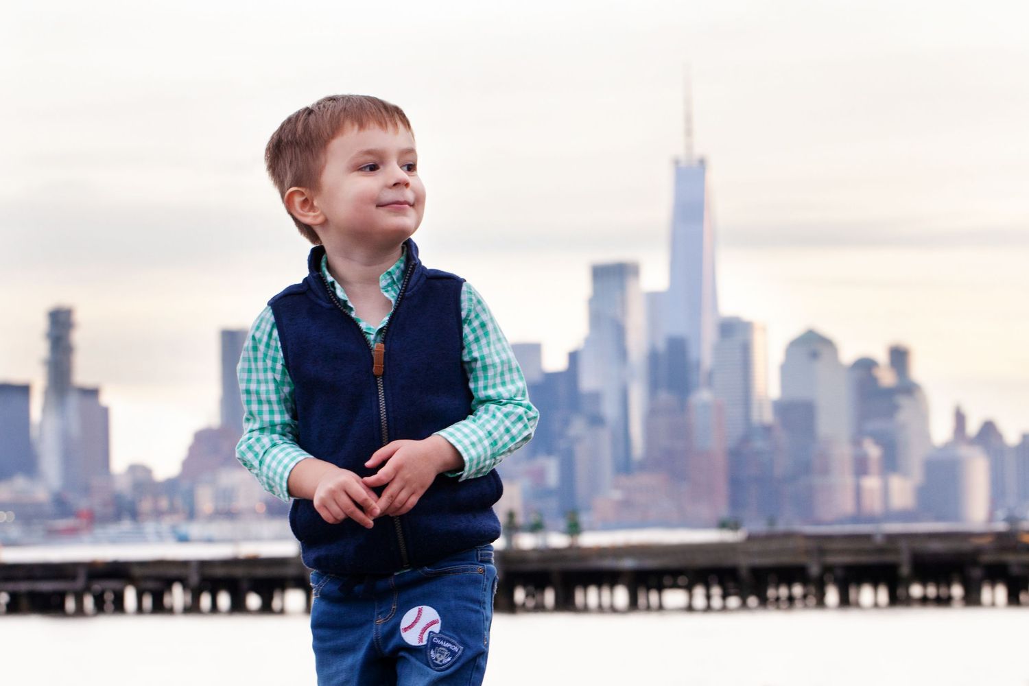 Young child in navy vest and checkered shirt stands with NYC Freedom Tower in background.