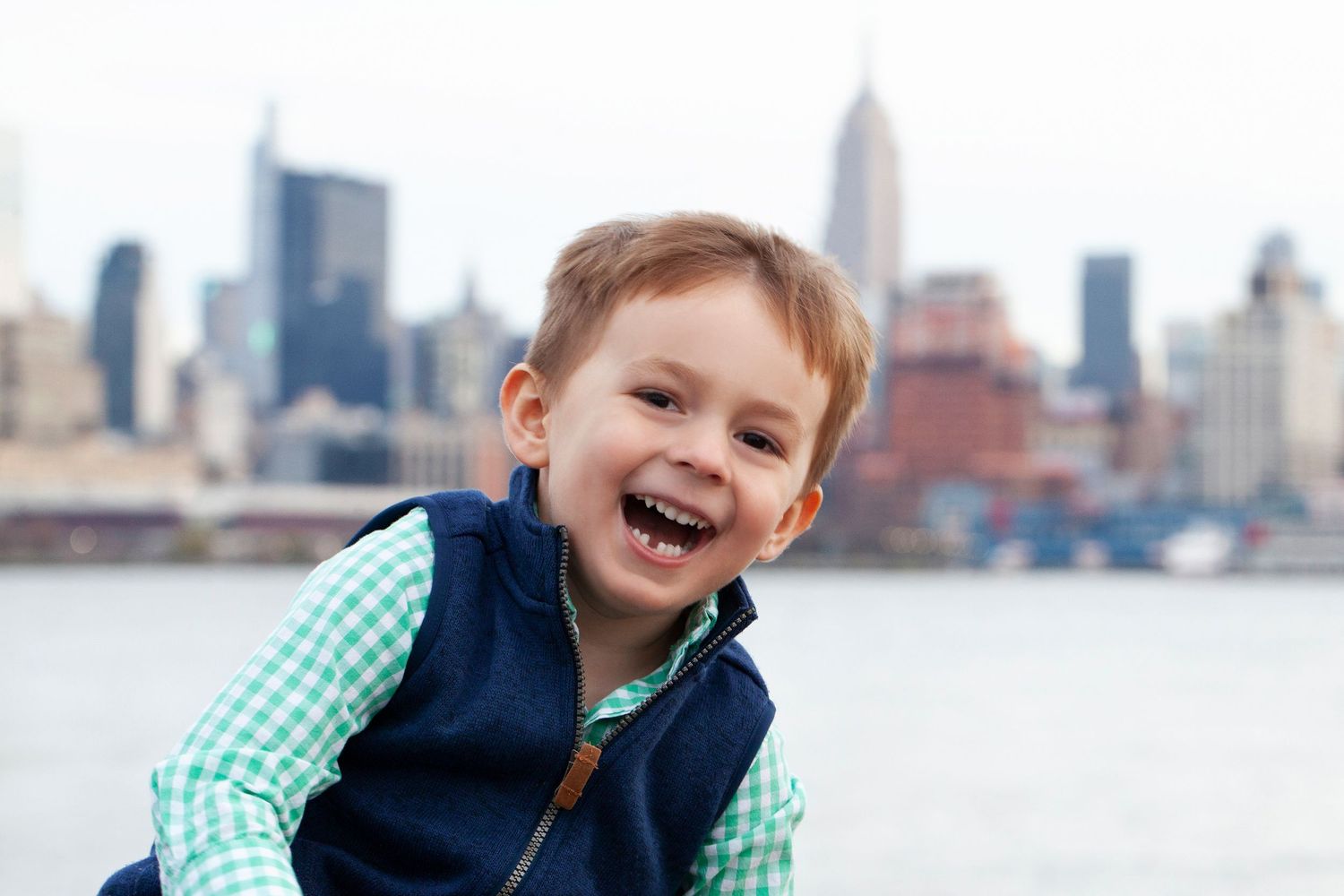 Cheerful kid in blue vest and gingham shirt poses against Manhattan skyline.