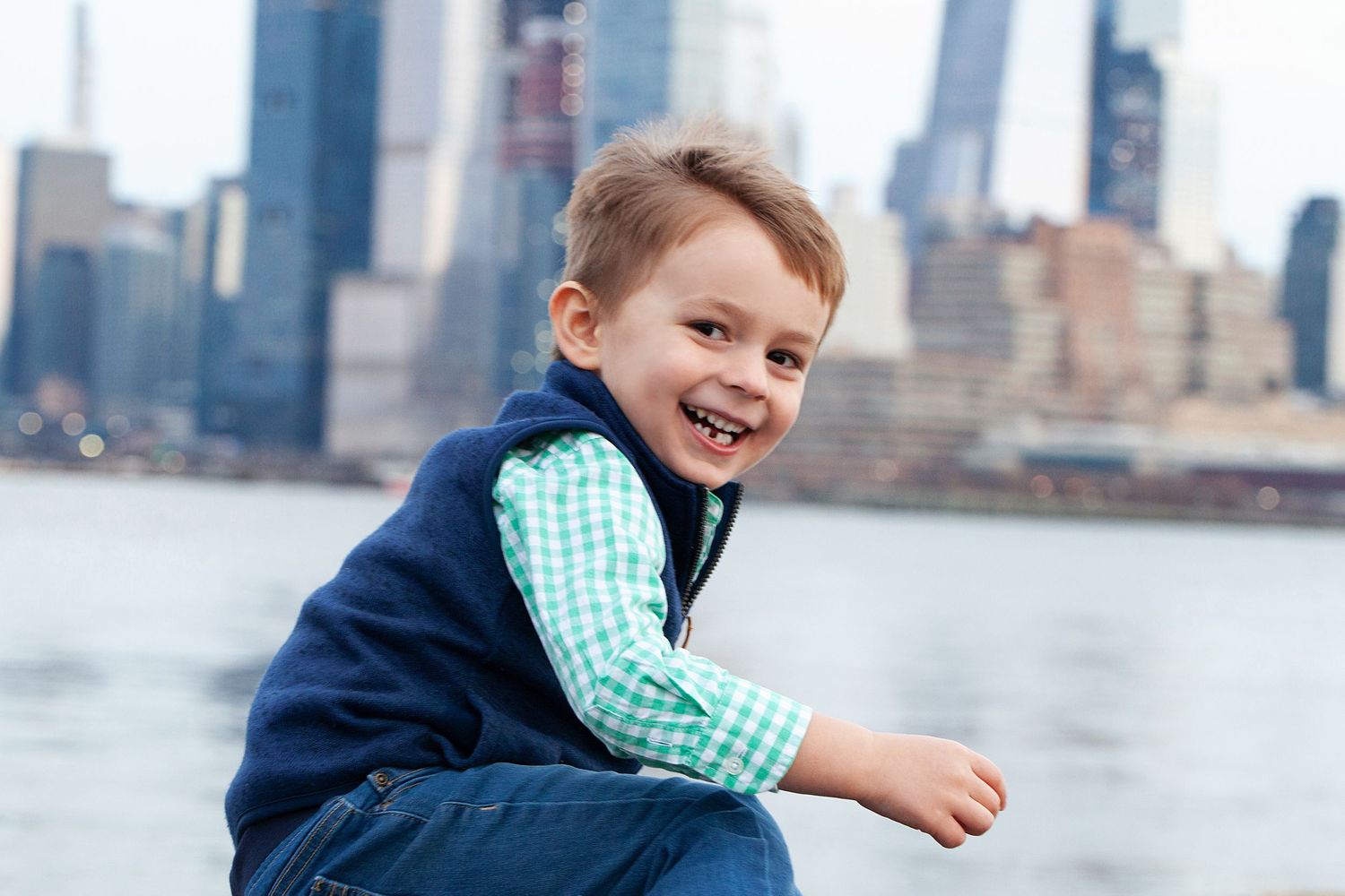 Child wearing blue vest and gingham shirt smiles while sitting by the New York City skyline.