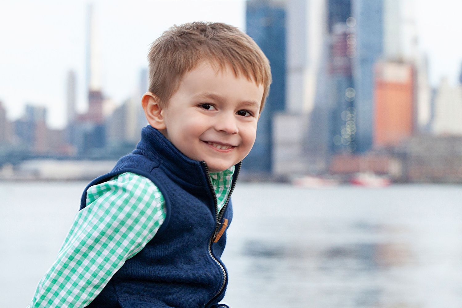Child wearing blue vest and gingham shirt smiling with Manhattan skyline in background.