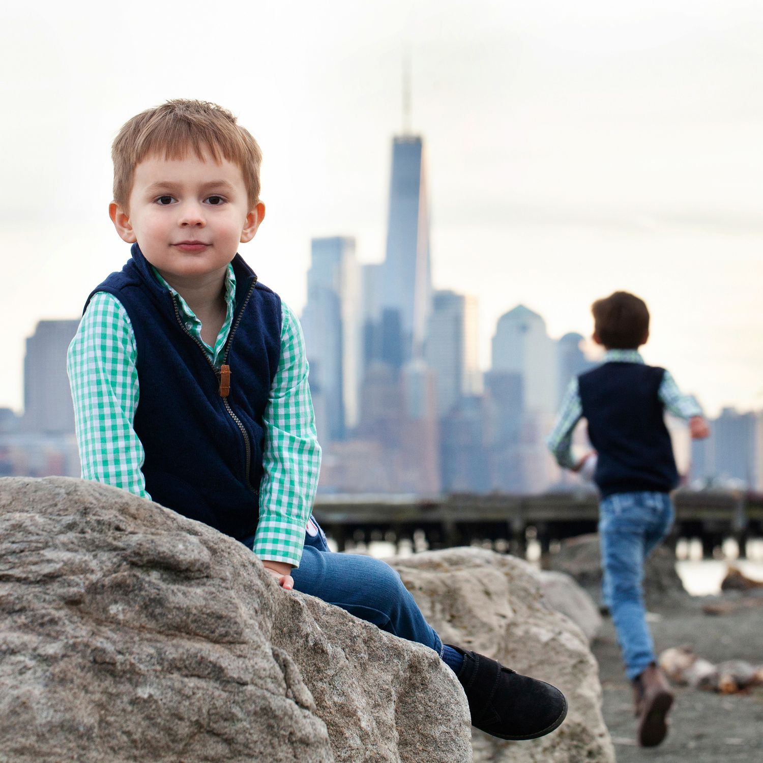 Child sits on rocky shoreline with Manhattan skyline and Freedom Tower in background.