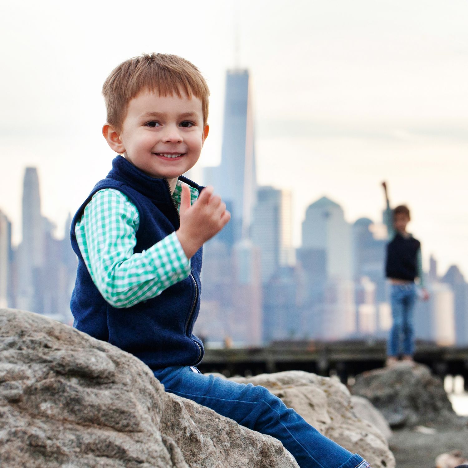 Child in blue vest sits on rocky shore while another figure stands in background near NYC skyline.