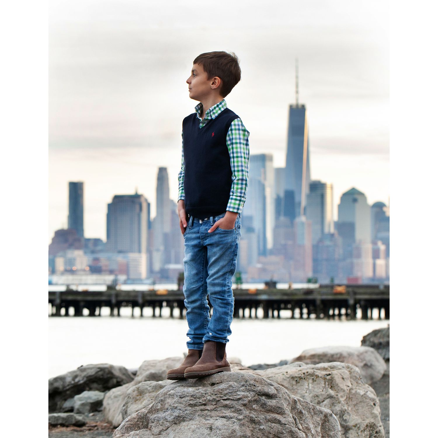 Child in navy vest stands confidently on rock with New York City skyline behind.
